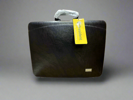 A black travel briefcase with a textured surface is shown, featuring a handle covered in plastic and a yellow tag attached to it. The case appears durable and has a sleek design suitable for professional use.